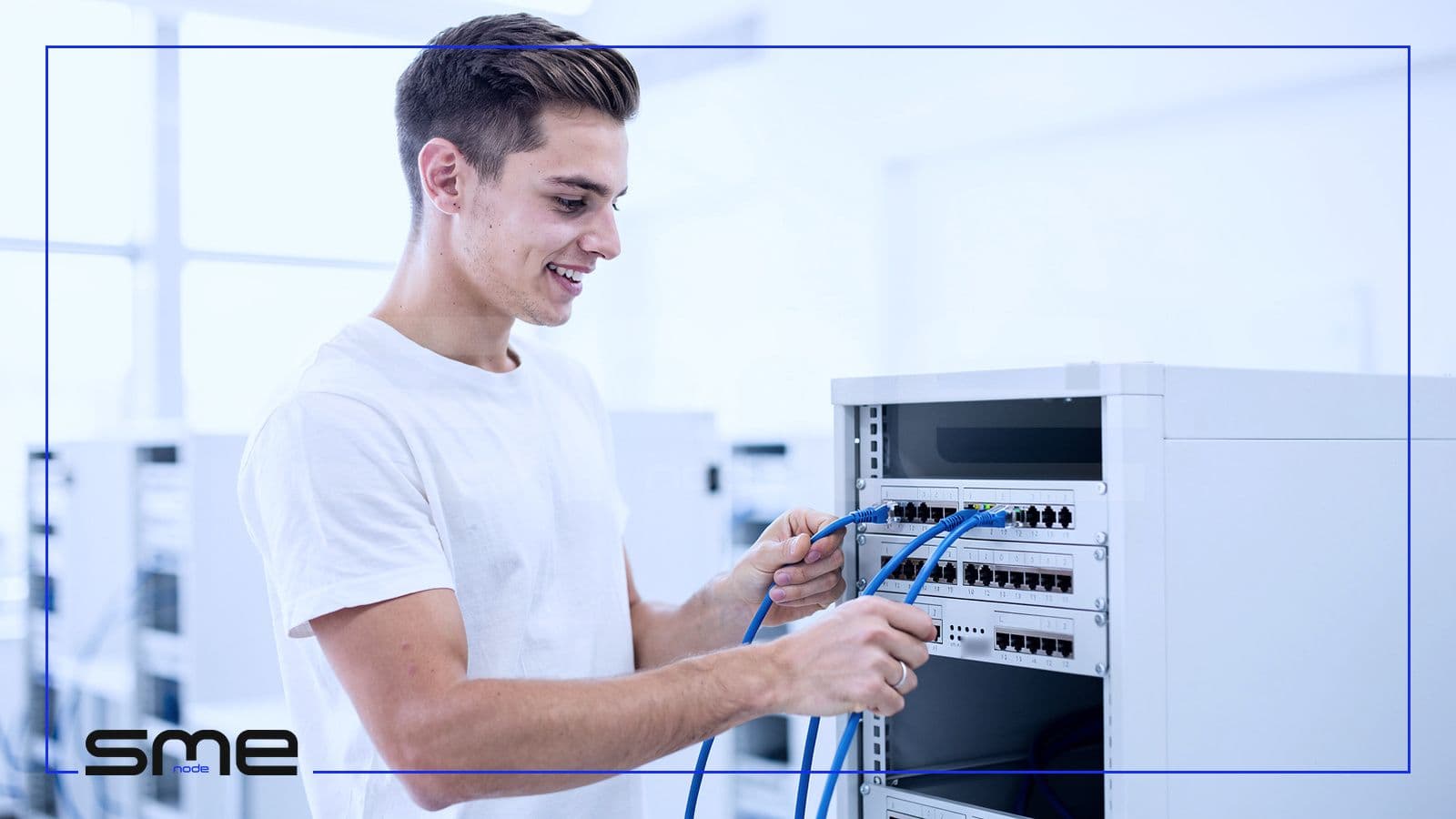 A young IT technician plugging blue patch cables into a network switch in a bright training lab