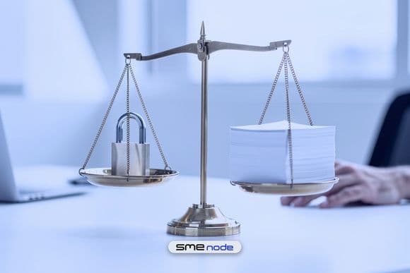 A brass balance scale on a white office desk holding a steel padlock on one side and a stack of blank paper on the other, level, to show security and rules weighed together in GRC