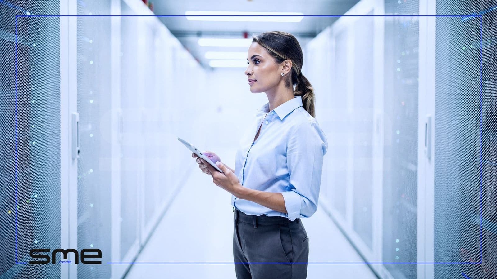 An engineer with a tablet standing in a bright data centre aisle between rows of server racks