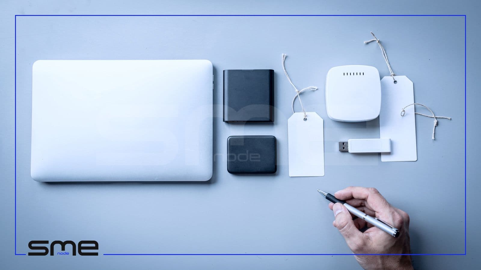Company devices laid out in a row on a grey table, a closed laptop, two external drives, a wireless router and a USB stick with blank paper tags, while a hand holds a pen ready to label them, the asset list that starts a risk assessment
