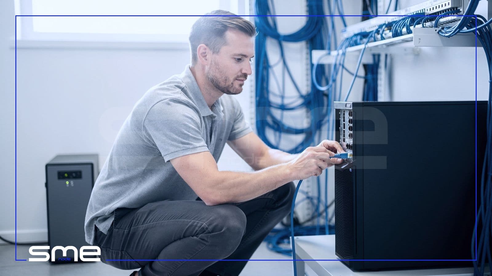 An IT technician plugging a network cable into a small on-premises tower server in a tidy office server closet with a UPS