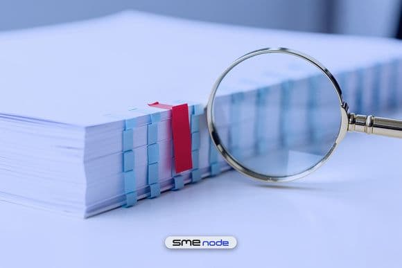 A thick white report with a row of pale blue index tabs and one red tab, beside a brass magnifying glass, showing how a reader hunts for the red flag in a vendor's SOC 2 report
